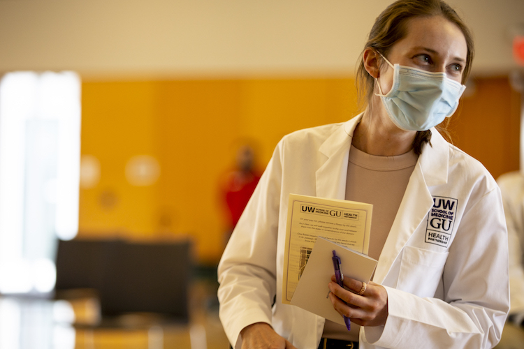 A person wearing a blue surgical mask and a white lab coat smiles at someone off-camera. They are holding paper and a pen.
