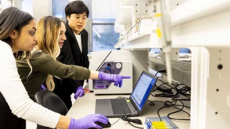 Three researchers discuss something on the screen of a laptop sitting on a lab counter
