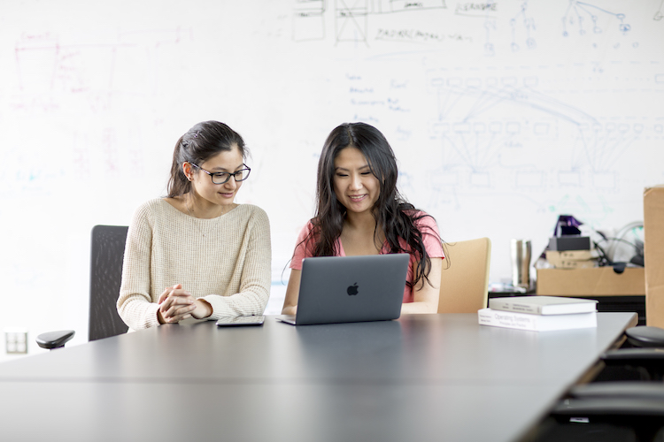 Two people sit at a metal table and look at a laptop computer. There is a whiteboard behind them covered in equations and diagrams written in blue ink.