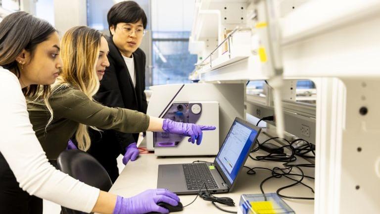 Three researchers discuss something on the screen of a laptop sitting on a lab counter