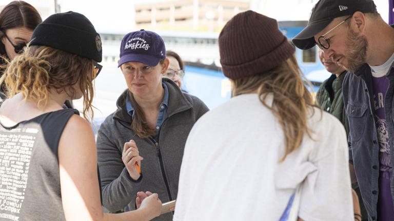A group of people look at a central item and at each other under a tent at the Fisherman's Terminal.
