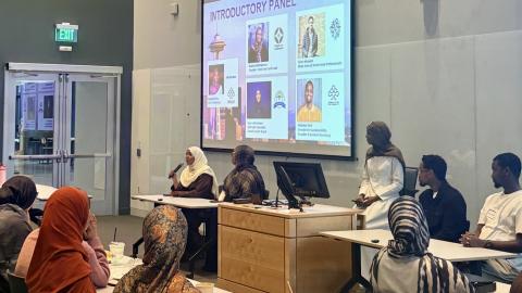 People gathered in a conference room with a large screen and panel of speakers