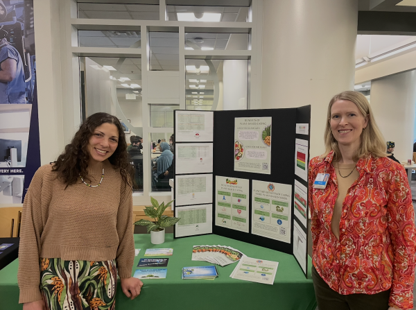 Two women smile and pose in front of a poster and table with educational materials