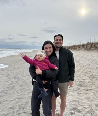 Two adults stand and pose with their young child on the beach